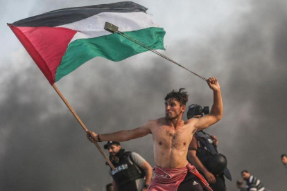 Un joven palestino tira piedras con una honda mientras con la otra mano lleva la bandera palestina.-MUSTAFA HASSONA / GETTY IMAGES