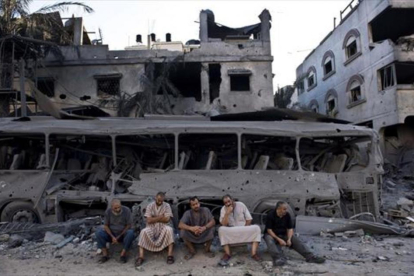 Cinco palestinos de Gaza sentados frente a un autobús y un edificio destruidos, en el 2014, por ataques aéreos israelís.-AFP / ROBERTO SCHMIDT