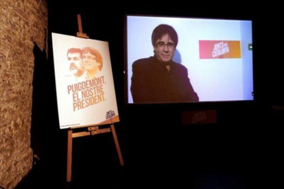 Videoconferencia de Puigdemont, junto a un cartel de Junts per Catalunya.-EFE / ALBERTO ESTÉVEZ