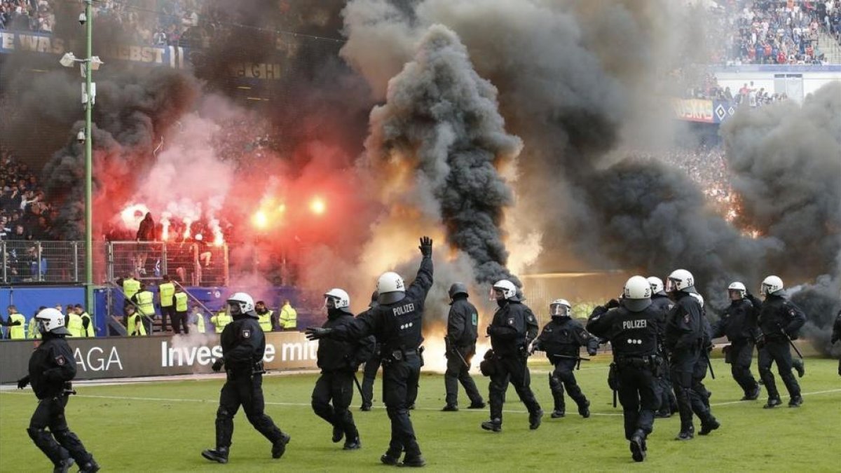 Antidisturbios saltan al césped del estadio del Hamburgo mientras en la grada se queman camisetas y banderas.-AP / MICHAEL SOHN