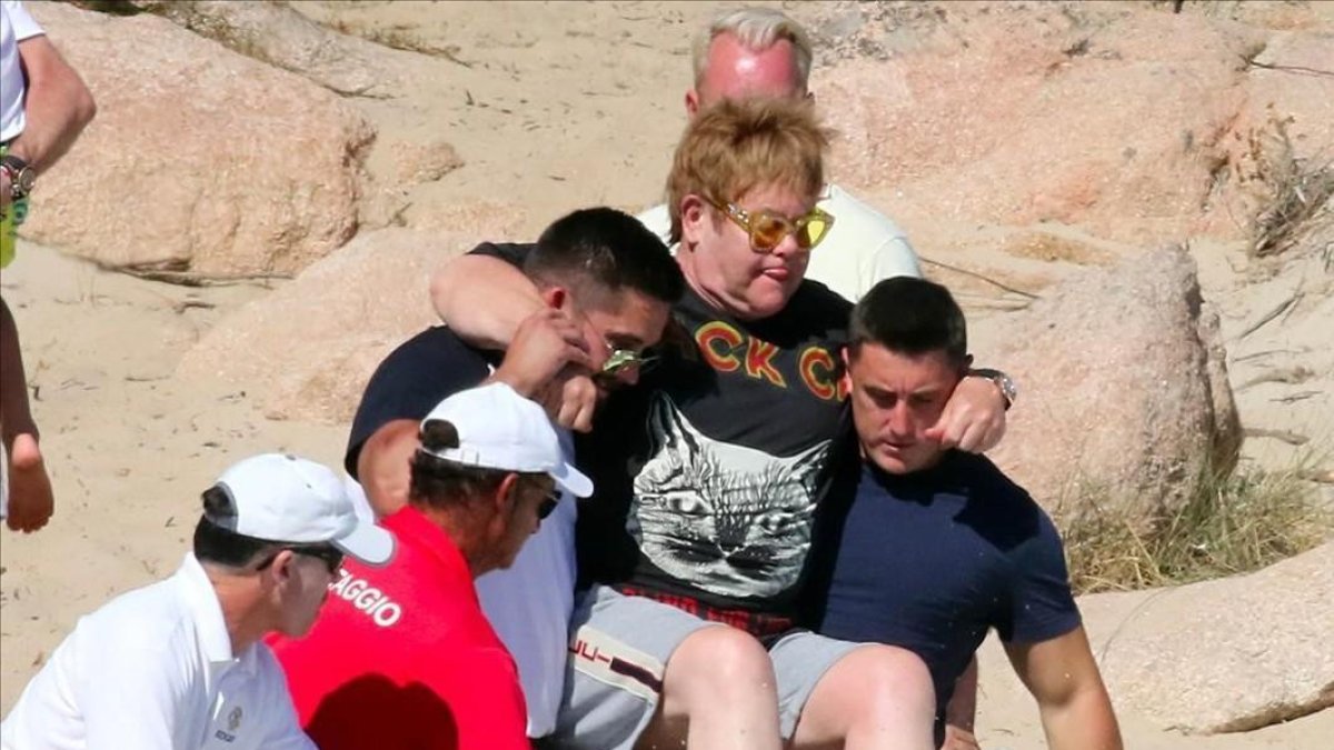 Elton John, cogido en volandas por sus guardaespaldas en la playa de Porto Cervo, en la isla de Cerdeña.-GTRES