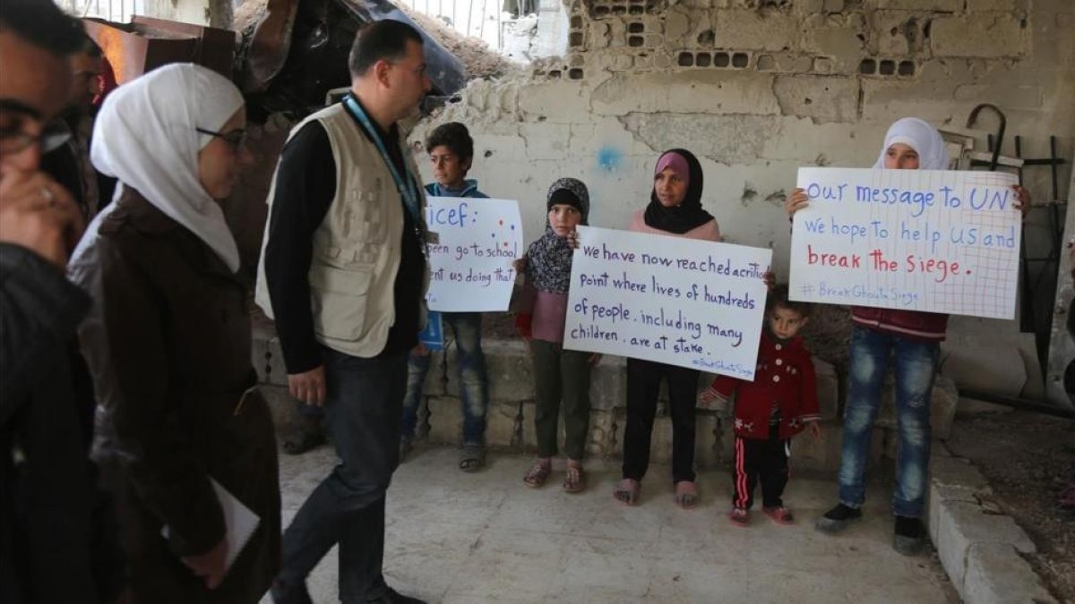 Niños sirios sostienen pancartas reivindicativas frente a miembros de un convoy de ayuda humanitaria durante una entrega de material en Al-Nashabia, en la región de Guta Oriental, al este de Damasco, el 28 de noviembre.-AFP / AMER ALMOHIBANY